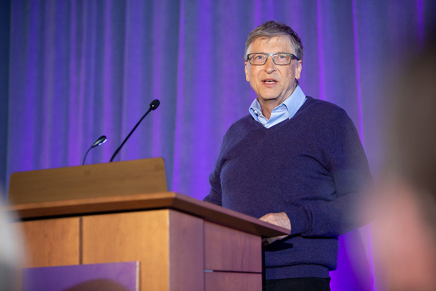 Bill Gates speaks at a podium against a purple backdrop.