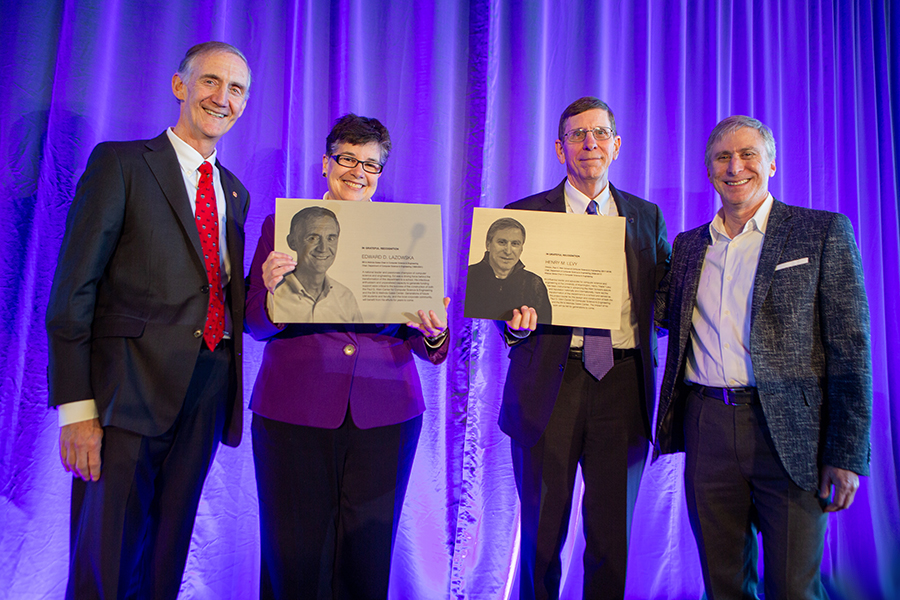 A row of four people in business attire standing onstage against a purple backdrop. The two people in the center are holding rectangular metal plaques depicting portraits of the other two.
