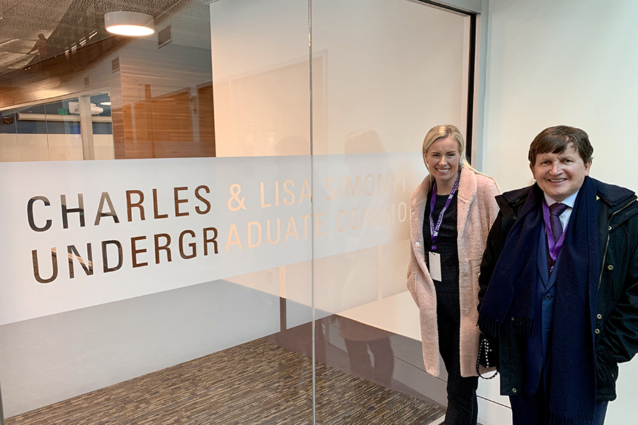 Two people standing outside of floor-to-ceiling glass doors with a white frosted sign on the glass that reads Charles & LIsa Simonyi Undergraduate Commons.