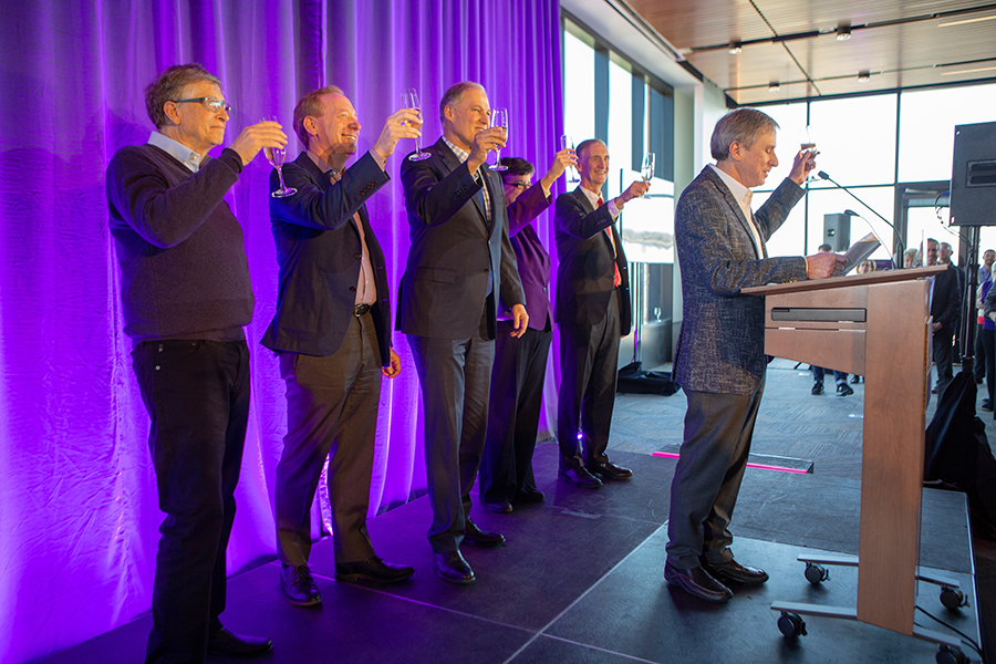 Hank Levy at the podium, with five people standing in a row behind, all raising their glasses in a toast against a purple backdrop.