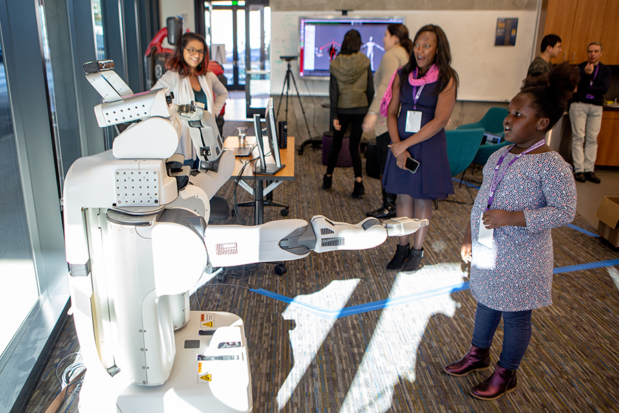 A girl looks with curiosity at a PR2 robot with its arm outstretched.