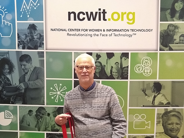 Richard Ladner seated on a red chair in front of NCWIT poster containing photos of people and line drawings in varying shades of blue and green