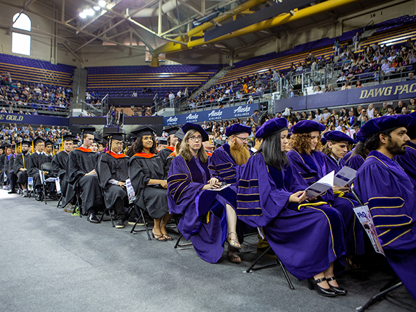 Graduates seated in the arena wearing caps and gowns, with guests seated in bleachers in the background