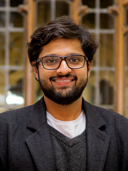 Dhruv Jain, wearing black glasses, a black sweater and a black blazer, smiles in front of a blurred background of windows.