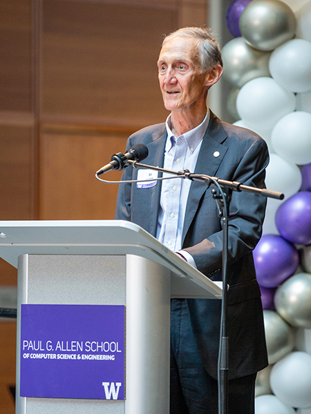Ed Lazowska stands speaking into a microphone attached to a silver podium with purple and white sign with text "Paul G. Allen School of Computer Science & Engineering" and UW block "W" logo. A column of purple, gold and white balloons is behind him off to the right. Wood paneling is visible in the background.