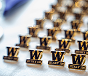 Display of gold and purple enamel lapel pins in the shape of "W" with "Husky 100" underneath on a white tablecloth