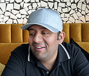 Venkatesh sitting at a black table with hands folded with a candid smile and a grey hat. The sofa is bright yellow and the wall is black and white pattern.