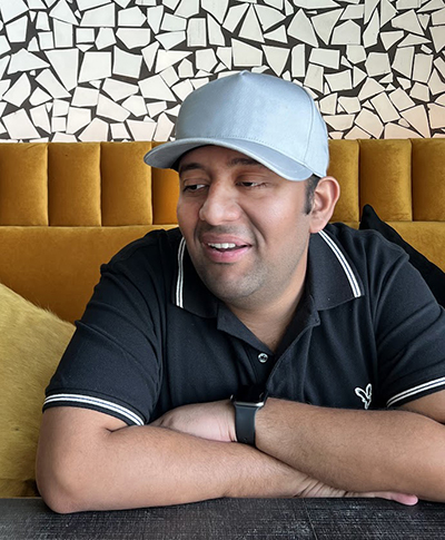 Venkatesh sitting at a black table with hands folded with a candid smile and a grey hat. The sofa is bright yellow and the wall is black and white pattern. 