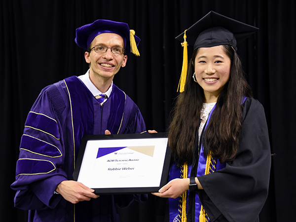 Robbie Weber and Lucy Jiang standing side by side onstage wearing graduation regalia and jointly holding a framed certificate.
