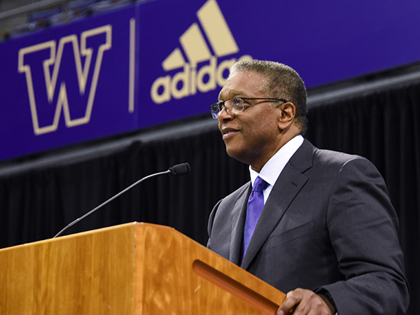 Side shot of Ron Howell wearing a dark suit and purple tie and glasses with his hand on the edge of a wood podium smiling behind a microphone. A purple banner with a gold outline of the UW block "W" logo and Adidas logo is in the background. 