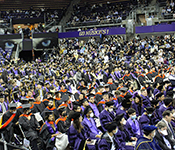 Wide shot of basketball arena full of people, with graduates in regalia seated in rows of chairs on the floor of the arena