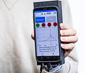 Hand holding smartphone attached to 3D-printed casing. Screen shows tympanometry software interface displaying status as "Measure" and peaking line graph.