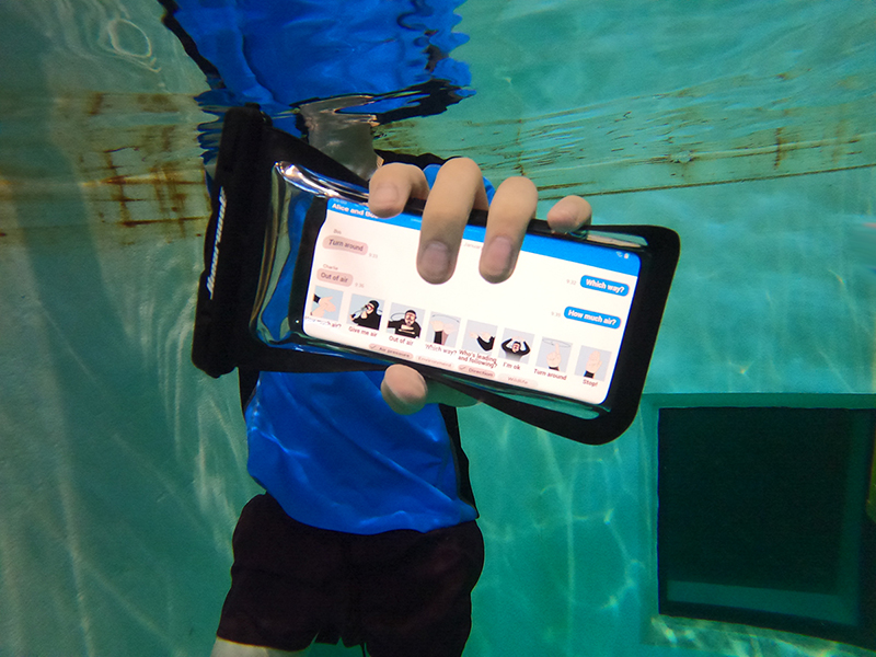 A person wearing a t-shirt and swim trunks holds a smartphone in flexible waterproof case underwater. The camera is focused on a close-up of the phone's screen showing the AquaApp interface with graphics depicting eight diving hand signals and a text exchange: "Which way?" "Turn around." "How much air?" "Out of air."