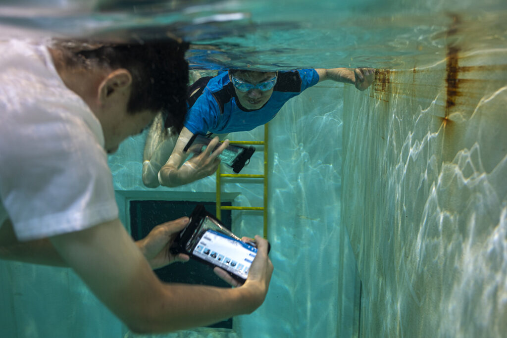 Two people in t-shirts and swimming trunks underwater in a tank holding smartphones in flexible waterproof cases. One of the smartphone screens is visible, displaying the AquaApp interface with text and graphics depicting various diving hand signals.