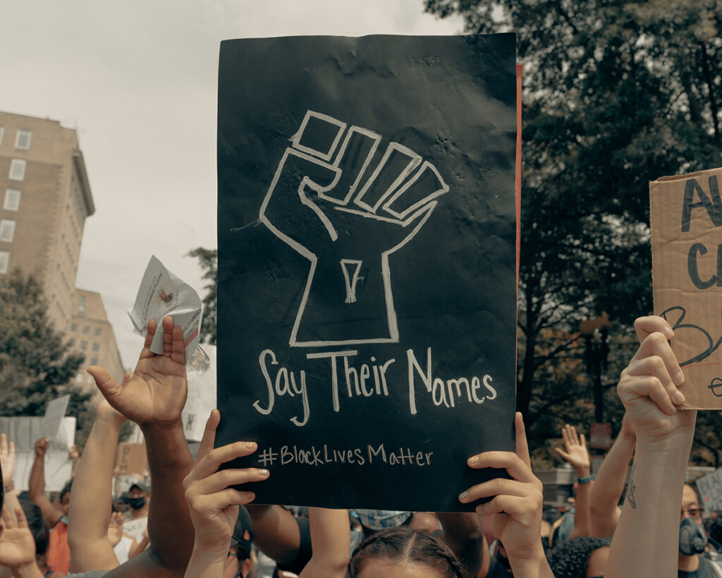 Protest participants marching toward camera with arms in the air, featuring a black sign with chalk drawing of raised fist and text "Say Their Names" and "#BlackLivesMatter" that one marcher is holding above their head. Only the forehead, hands and wrists of the sign holder is visible.