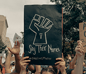 Protest participants marching toward camera with arms in the air, featuring a black sign with chalk drawing of raised fist and text "Say Their Names" and "#BlackLivesMatter" that one marcher is holding above their head. Only the forehead, hands and wrists of the sign holder is visible.