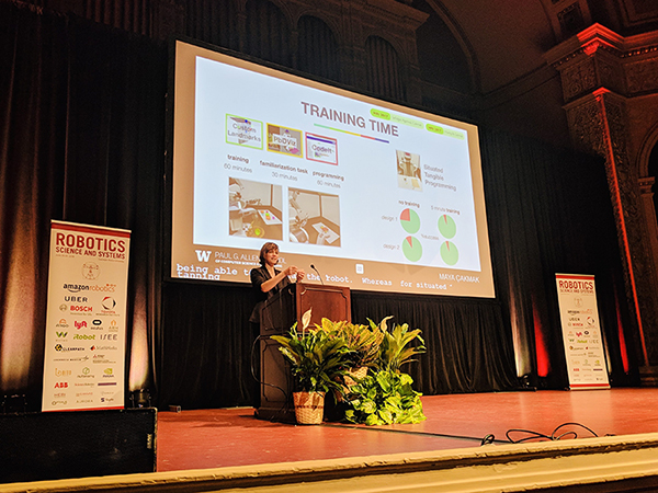 Maya Cakmak standing onstage behind a solid wood podium with assortment of potted foliage in front, talking into a microphone. There is a screen behind her with a PowerPoint slide titled "Training Time" with assorted images and charts, flanked by two signs with text "Robotics Science and Systems" and multiple company logos underneath.