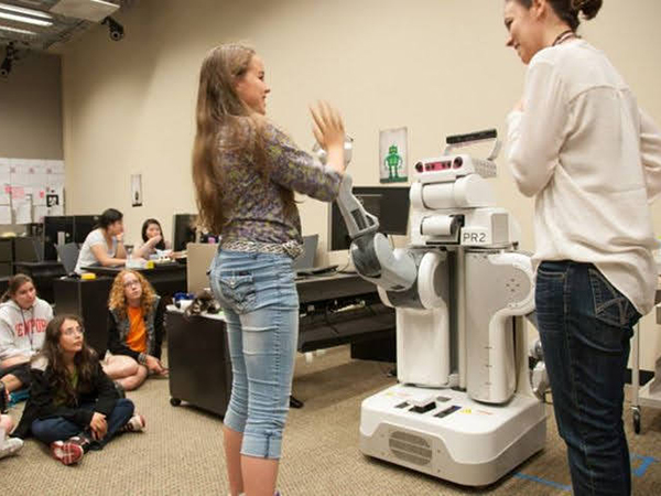 Maya Cakmak stands smiling at a girl who appears to be middle-school age. The girl is smiling back while holding her right hand up towards a PR2 robot, which has its right hand raised, as a group of other girls sitting cross-legged on the floor looks on. There are desks with large computer monitors on them lining the wall behind them.