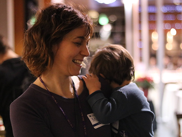 Maya Cakmak in profile, smiling and holding a baby facing away from the camera, with a blurred Allen Center atrium at night in the background.