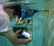 Two people in t-shirts and swimming trunks underwater in a tank holding smartphones in flexible waterproof cases. One of the smartphone screens is visible, displaying the AquaApp interface with text and graphics depicting various diving hand signals.
