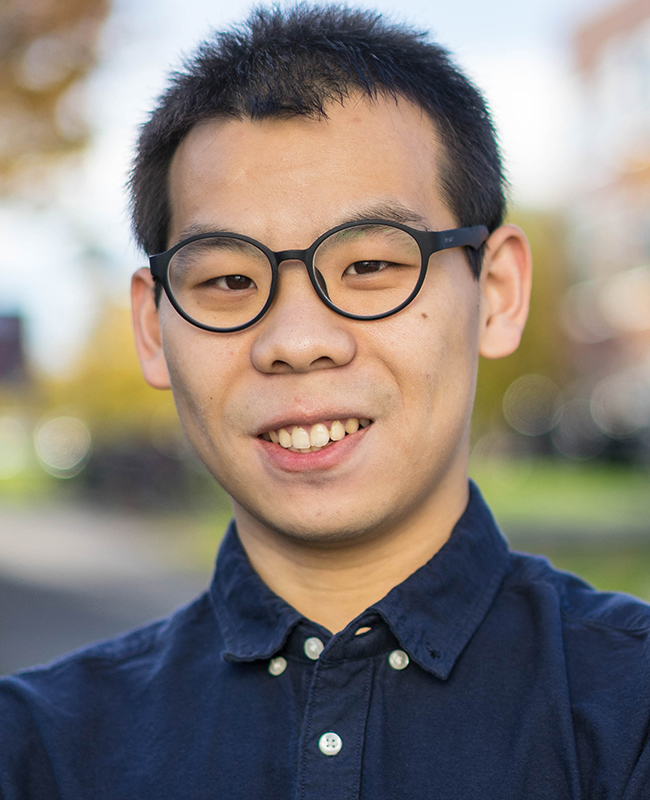 Portrait of Xin Liu in dark blue button-up shirt and glasses standing outdoors with fall foliage and buildings blurred in the background.