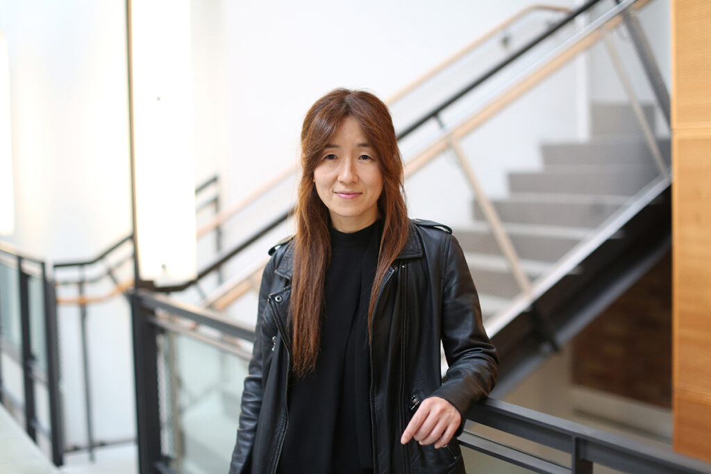 Yejin Choi in a black leather jacket over a black shirt, leaning against a metal railing with a metal, wood and concrete stairwell in the background. A portion of wood-paneled wall is visible in the right of the frame.