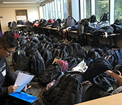 Student backpacks of various colors and styles piled on tables against a wall of glass windows and on the floor. A person wearing glasses and a shirt printed with the Allen School name and a stick-on name tag is crouched in the lower left corner of the photo, inserting papers into a blue plastic folder