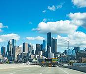 Seattle skyline viewed from a three-lane highway framed by street lamps, with vivid blue sky and fluffy clouds