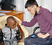 ALT TEXT: A young boy wearing a grey and black zip-up jacket with green and orange trim and jeans sits in an ivory-colored plastic chair as a researcher seated adjacent to him in a dark grey office chair holds a probe to his ear. The probe is attached to a smartphone sitting on the researcher's lap. The researcher is wearing glasses, with a mask around his chin, a plaid button down shirt and dark cotton trousers. The child is looking down at the smartphone screen. There is a desk with papers and a pen and a backpack behind the child.