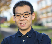 Portrait of Xin Liu in dark blue button-up shirt and glasses standing outdoors with fall foliage and buildings blurred in the background.