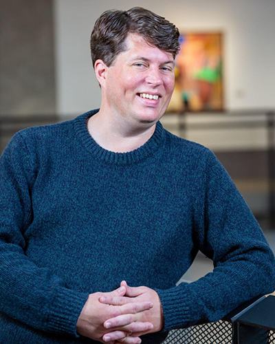 Joe Eckert, wearing a dark blue sweater, leans on a railing while smiling in front of a blurred background, possibly a painting hung on a wall.