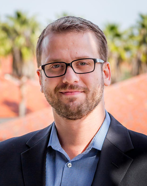 Portrait of Baris Kasikci with short, light-brown hair and trim facial hair wearing glasses and a medium-blue button-down shirt under a dark suit jacket. Red tile roofs and palm or possibly banana trees are blurred but visible in the background.