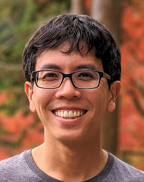Portrait of Pang Wei Koh with short tousled dark hair wearing glasses and a grey t-shirt. Trees with autumn-colored leaves and evergreens are blurred but visible in the background.