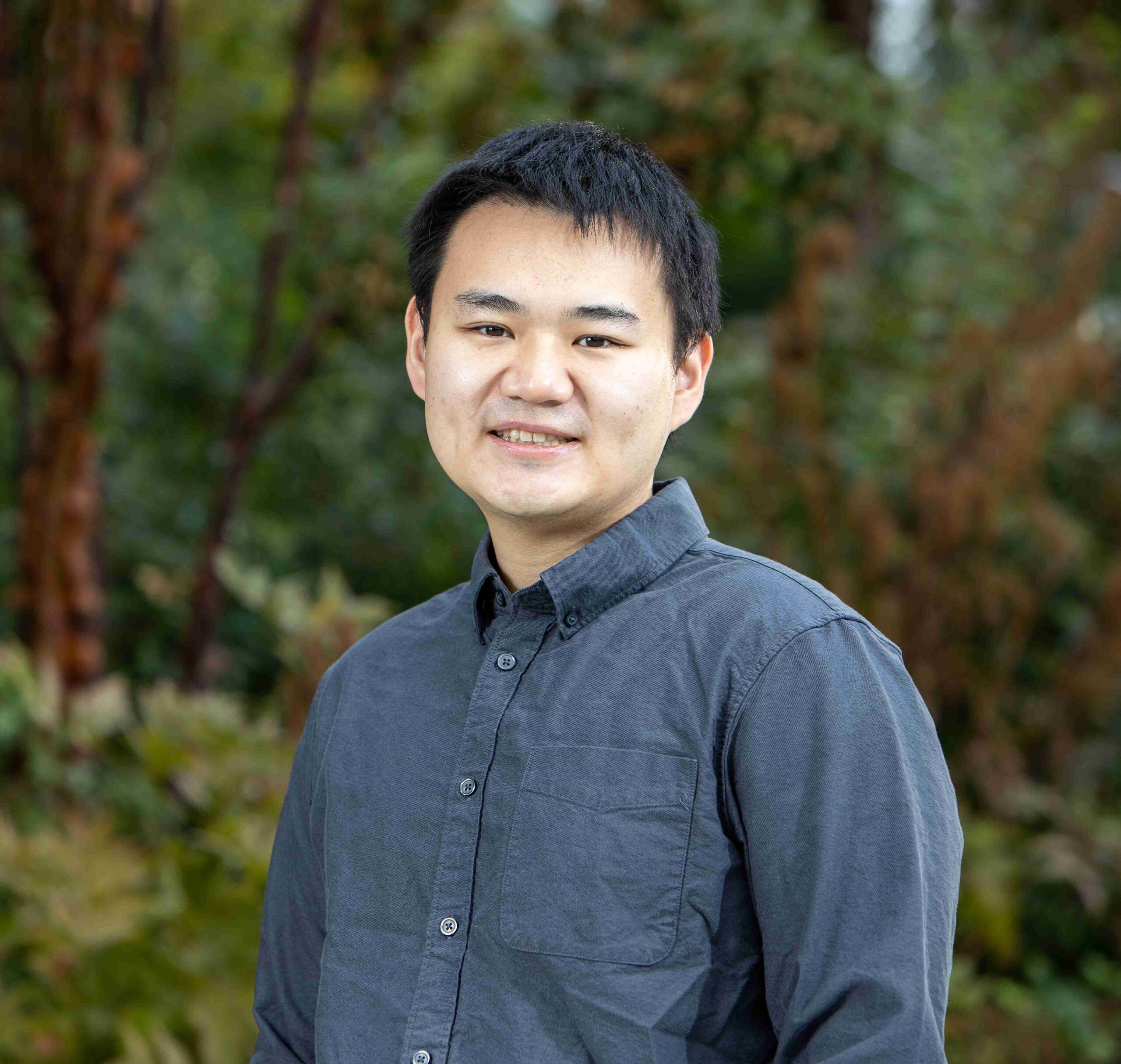 Simon Du, wearing a navy blue shirt, smiles while standing in front of a wooded background.