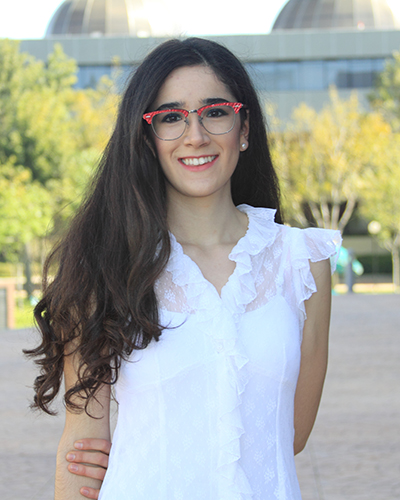 Sofia Padilla Munoz stands in front of a gray building with two domes and there are trees in front of the building. She is wearing a white shirt and red glasses.