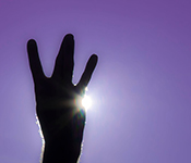 Hand silhouetted against the sun, giving the "dubs up" symbol, with ring and middle finger crossed in between pinky and index finger to resemble the letter "W," against a purple background