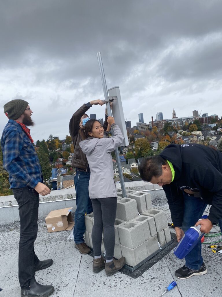Four volunteers from the Seattle Connectivity Lab install a rooftop station consisting of a rectangular metal piece roughly two feet high on a thin metal pole, anchored in a pile of cinderblocks, with cityscape and cloudy sky in the background. One person is holding the station in place while the other secures the metal box to the pole, while a third person looks on and a fourth person squats nearby and appears to be gathering tools.
