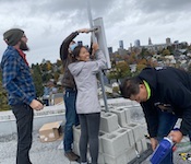 Four volunteers from the Seattle Connectivity Lab install a rooftop station with city scape and cloudy sky in back drop.