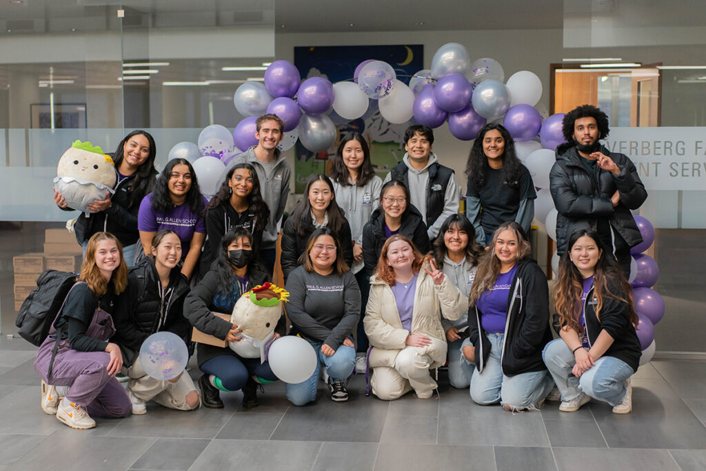 Group photo of 18 people, some of whom are wearing Allen School-branded clothing, framed by a purple, silver and white balloon arch in front of glass doors inside the Gates Center atrium. Two people are holding round white stuffed animals resembling white blobs with black eyes and felt hats, and two people are holding individual balloons.