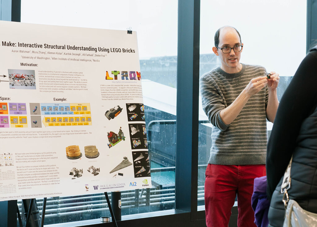 A person wearing glasses, a grey and white striped sweater, and red trousers is holding a small item, possibly a LEGO piece, in his hands and talking to a person who is positioned with their back to camera, mostly out of frame. Next to the person talking is a poster with colorful images and diagrams of various Lego building projects, with a partially visible title text: “Make: Interactive Structural Understanding Using LEGO Bricks.” The poster is in front of a large glass window overlooking the exterior of Husky Stadium in the rain.
