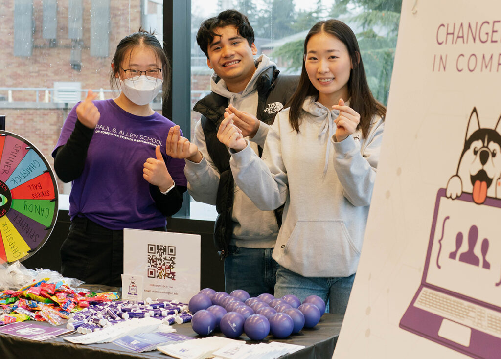 Three people, one of whom is wearing glasses, a purple Allen School t-shirt and a mask and the others wearing light grey sweatshirts, one with a North Face puffer vest on top, standing behind a table making snapping gestures with their fingers and smiling at the camera. Part of the “Changemakers in Computing” sign with a graphic of a Husky and laptop with three avatars in brackets onscreen is visible. There are piles of purple rubber balls and chapstick tubes, a selection of purple and white stickers with Allen School and Changemakers in Computing’s Husky logo, and a pile of candy behind a multi-colored game wheel that people can spin to win items on the table. A QR code in a plastic sheath is also on the table. Evergreen branches and a brick building are visible through the window in the background.
