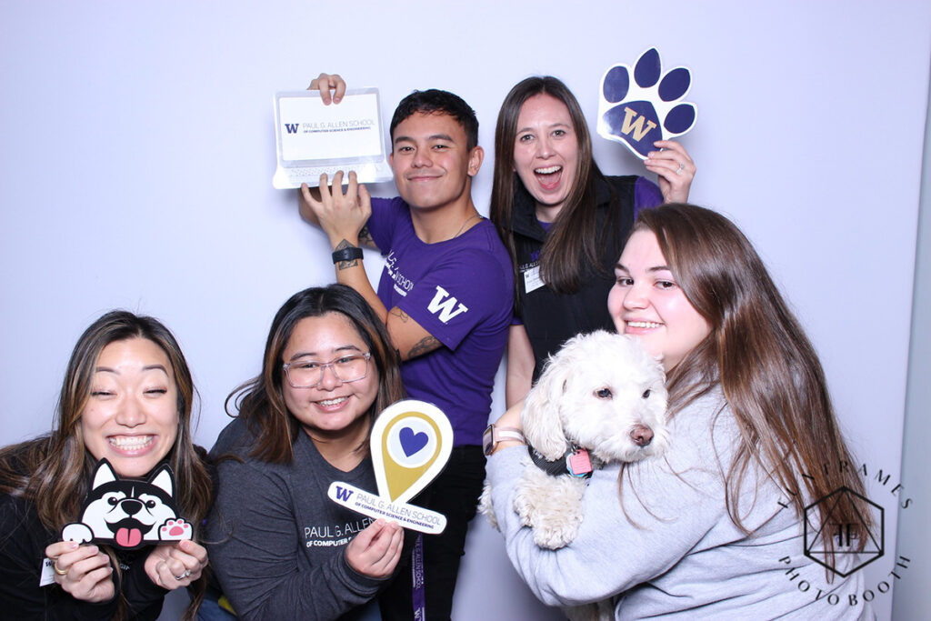 Five smiling people, one of whom is holding a dog with curly white hair and pink and brown nose, pose in a photo booth. Two of the people are wearing t-shirts with UW and Allen School logos visible. The four people without the dog are holding up 2D props, including a graphic of a Husky with pink tongue and paw pads, an online map-style location pin with a heart in the center and the Allen School logo underneath, an open laptop with the Allen School logo onscreen, and a purple dog paw with a gold “W” on the paw pad.