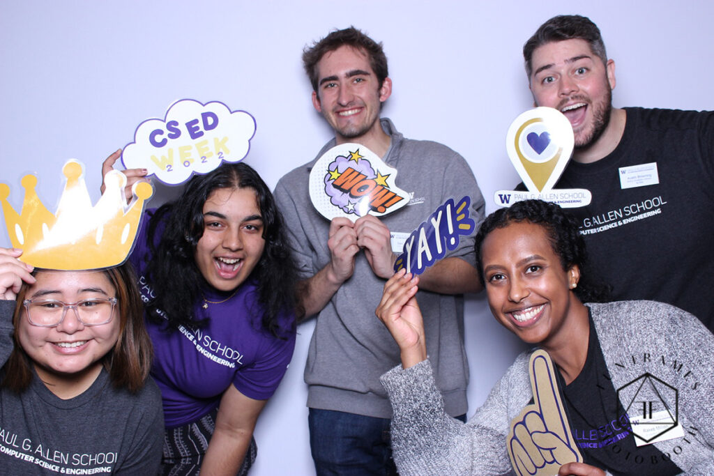 Five people, four of whom are wearing Paul G. Allen School of Computer Science & Engineering t-shirts, pose with 2D props in a photo booth, including a sign shaped like a crown, a thought cloud with the text CS Ed Week 2022, a comic book-style graphic with stars and the text WOW!, an online map-style location pin with a heart in the center and the Allen School logo underneath, a cut-out of the word YAY!, and a sign shaped like a hand with index finger pointing up in a “#1” gesture.
