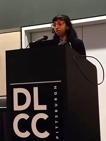Cailin Winston stands behind a podium and laptop, speaking into the connected microphone. The podium is black with white text "DLCC PITTSBURGH," and a pair of light-colored doors and two-toned walls of the banquet room, in sage green and cream, are visible behind her.