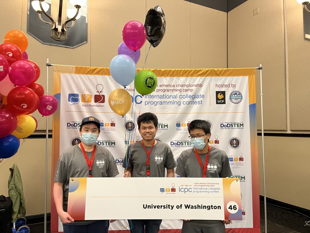 Nathan Akkaraphab, Milin Kodnongbua, and Phawin Prongpaophan stand in front of a white banner while holding a placard with the words, the University of Washington, on the front. Kodnonbua holds the strings of several multicolored balloons. 