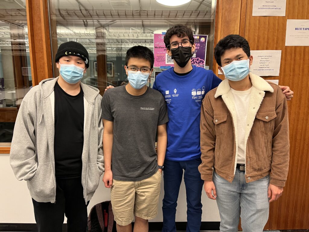 Nathan Akkaraphab, Phawin Prongpaophan and Milin Kodnongbua stand shoulder-to-shoulder with Victor Reis, who is wearing a blue shirt and glasses. Each is wearing a mask. They are standing in front of a window and a wooden door. 