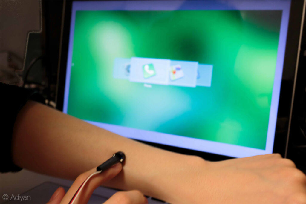 Person sliding fingertip with small sensor device across their forearm in front of a computer screen showing a selection of menu icons superimposed on each other horizontally, with the green and white phone icon selected