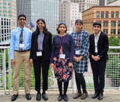 Group photo of the five Winston siblings wearing business attire and name tags on lanyards around their necks, standing side-by-side in front of a metal railing with buildings of various styles of stone, metal and glass in downtown Pittsburgh in the background.