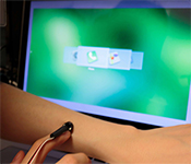 Person sliding fingertip with small sensor device across their forearm in front of a computer screen showing a selection of menu icons superimposed on each other horizontally, with the green and white phone icon selected