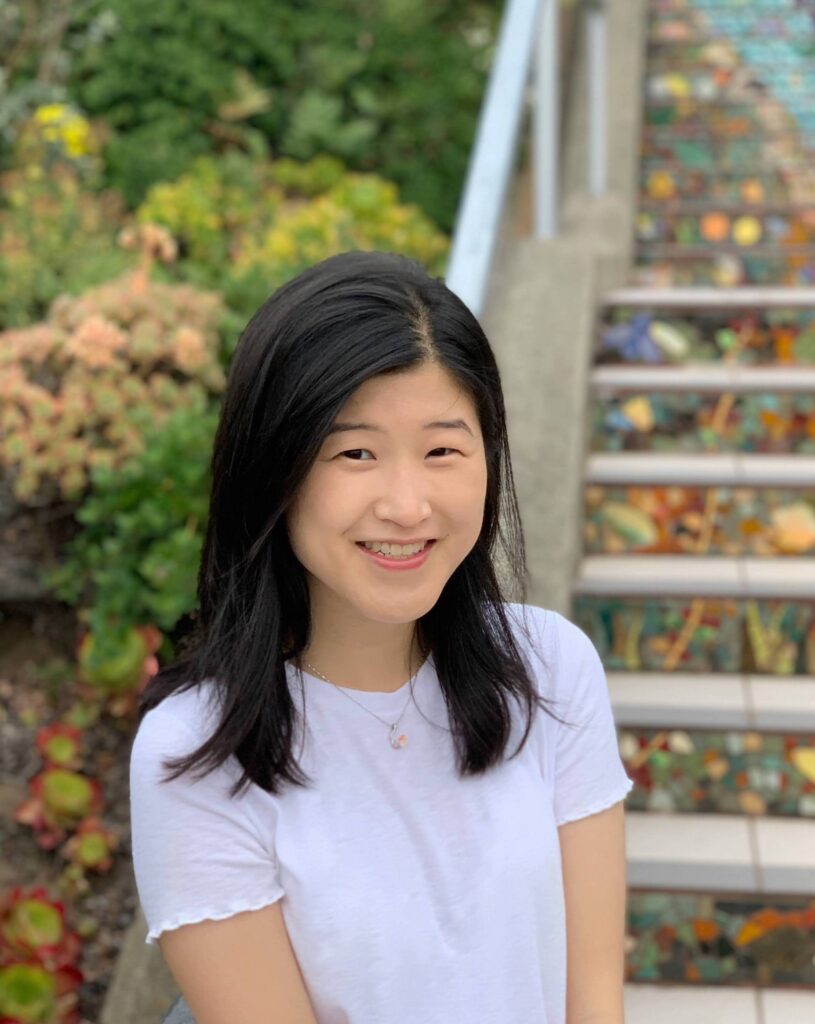 Portrait of Alisa Liu wearing a white short-sleeved shirt with gathered short sleeves and a pendant necklace standing in front of a mosaic tiled staircase and foliage of succulents, ferns, and bushes.